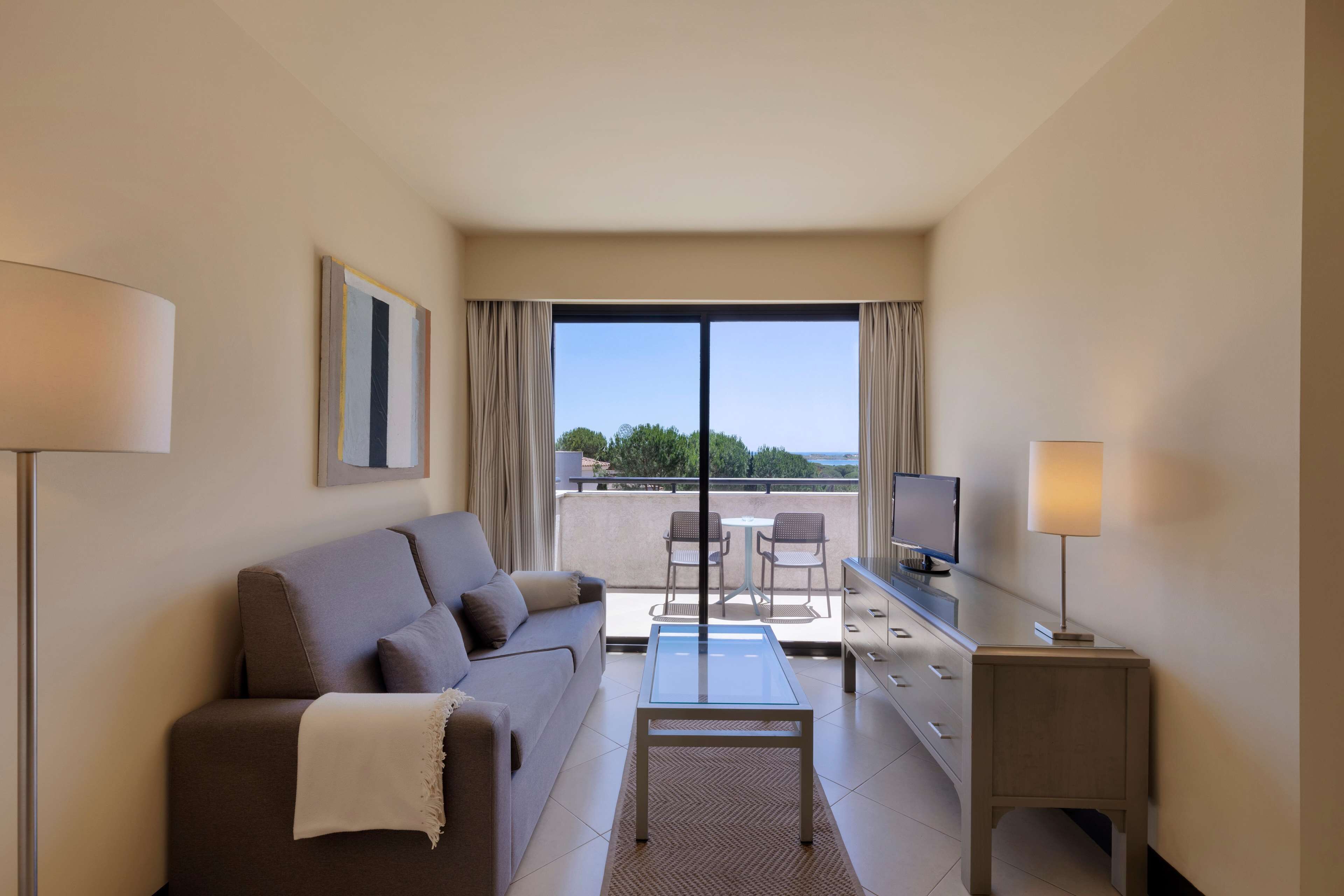 A modern hotel room with a sofa, a glass coffee table, a desk with a lamp, a TV, and a balcony with chairs.