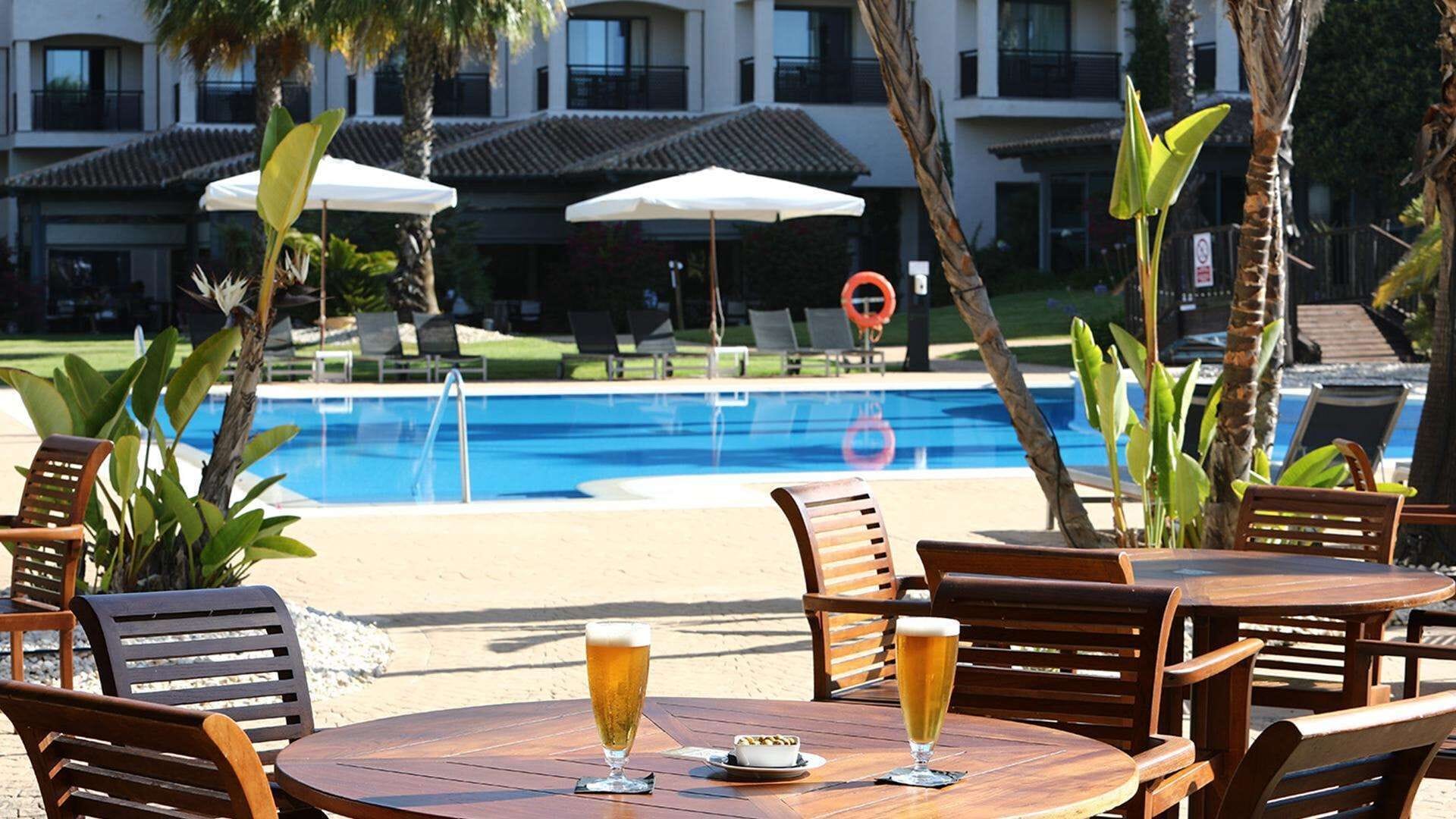 Zona junto a la piscina con dos mesas redondas de madera, cada una con una cerveza y un cuenco pequeño de frutos secos. Se ven sombrillas, sillones de descanso y un edificio con balcones al fondo.