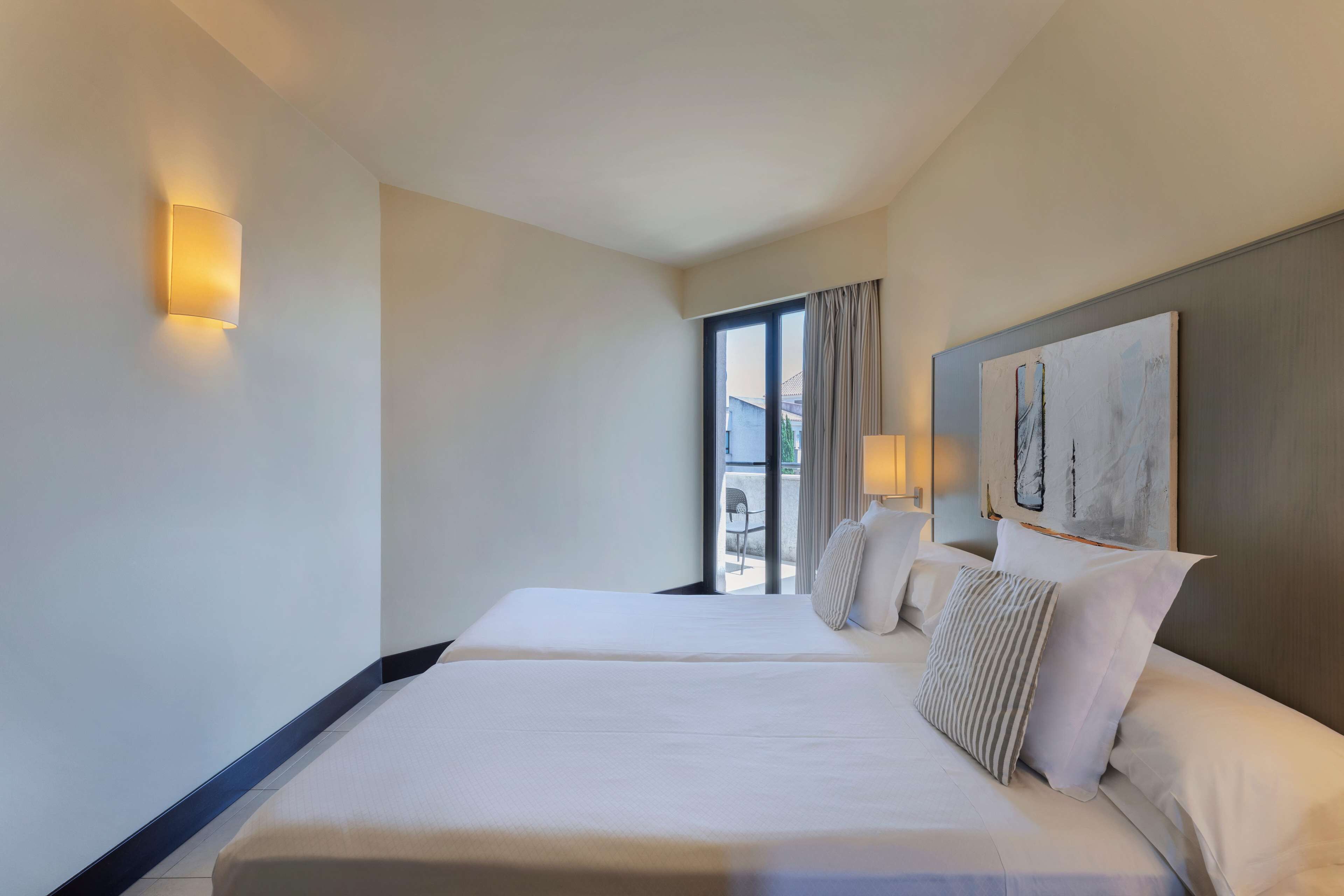Habitación de hotel con dos camas, un ventanal con cortinas, un cuadro en la pared y una lámpara fijada a la pared.