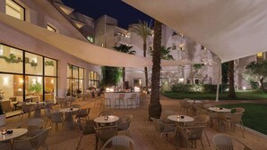 An outdoor dining area with multiple tables and chairs, a bar, and a building with large windows and plants.