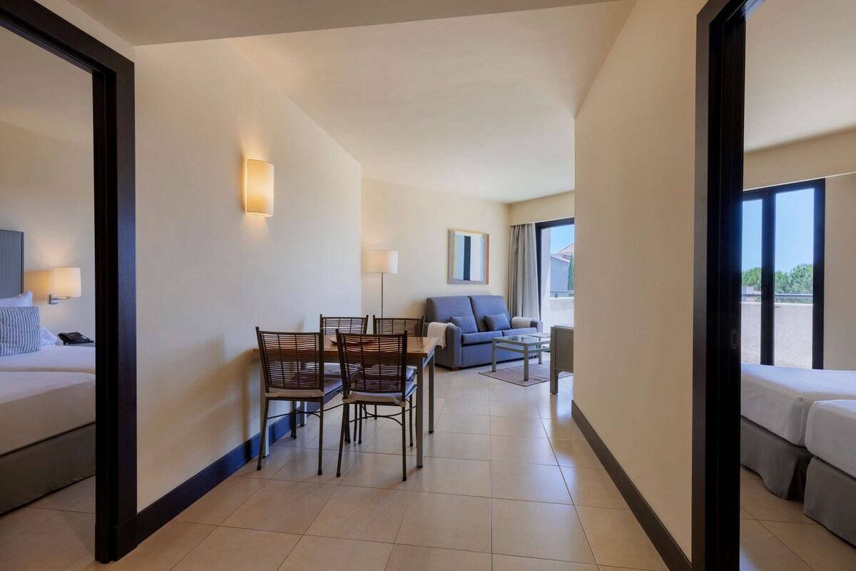 A hotel room with a bed, a dining table with chairs, a sofa, and a large window.
