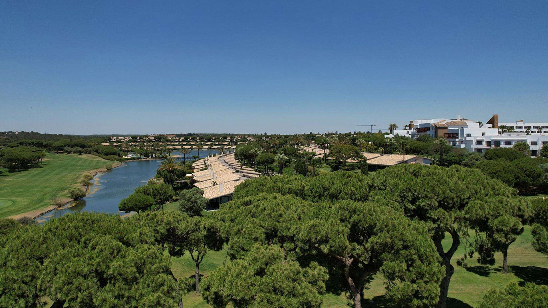Un campo de golf con un río, rodeado de abundante vegetación y edificios modernos.