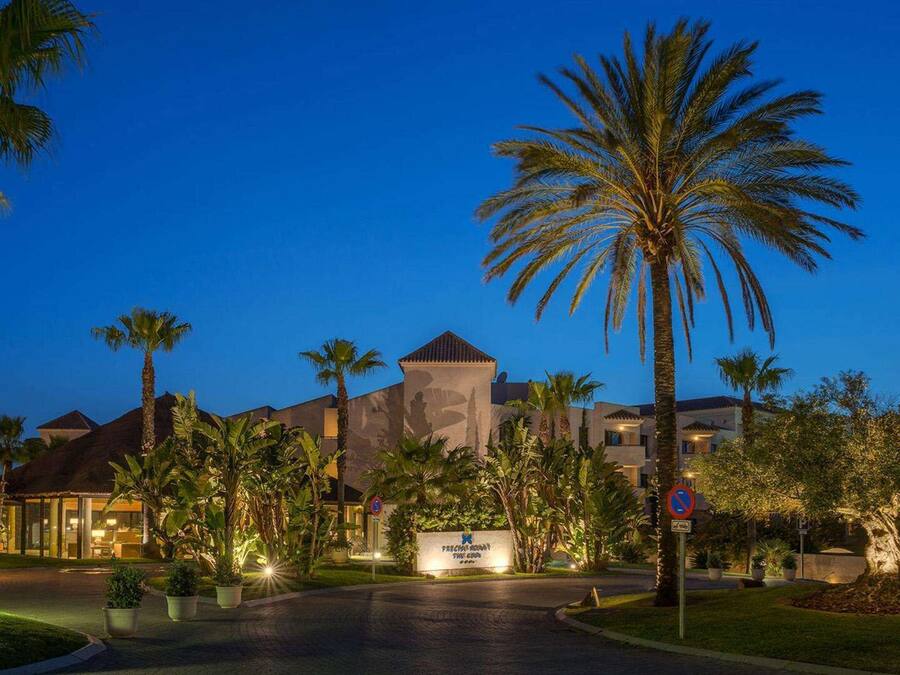 A hotel with a central building, palm trees, and a well-lit entrance.