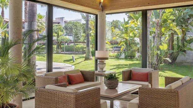 A sunlit room with wicker furniture, a table, and a view of a lush garden.