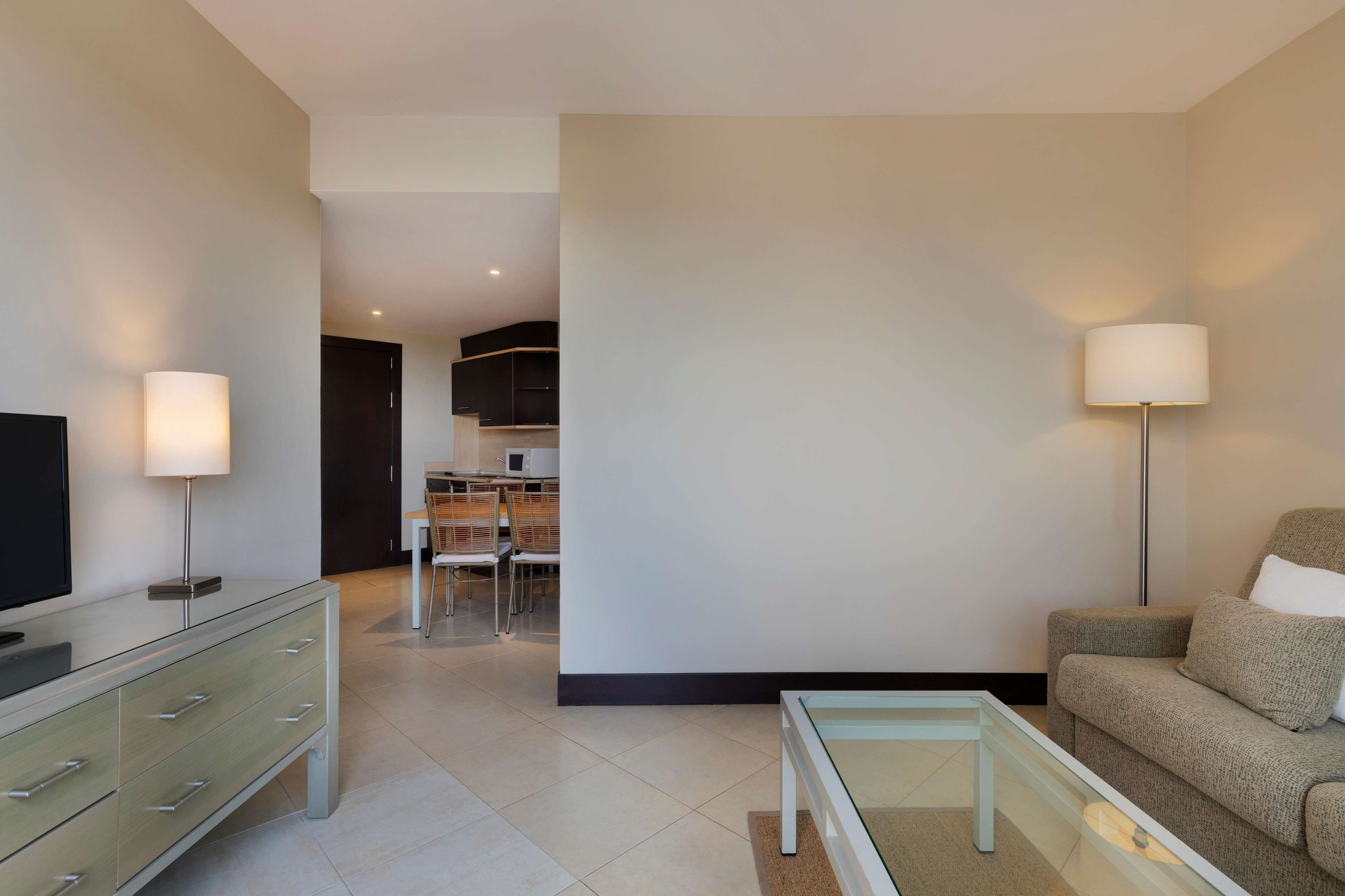 Una sala de estar moderna con un sofá, una mesa de centro de vidrio, un televisor sobre un mueble bajo y un área de comedor al fondo.