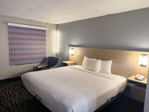 Ein Hotelzimmer mit einem großen Bett, einem Schreibtisch, einem Stuhl, einem Nachttisch mit Telefon und einem gemusterten Fensterrollo.