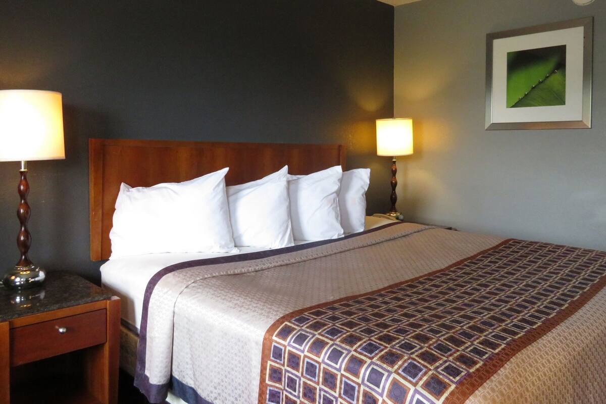 Ein Hotelzimmer mit einem hölzernen Kopfteil, einem Bett mit weißen Leinen, zwei Nachttischlampen, einem Nachttisch und einem gerahmten Bild an der Wand.