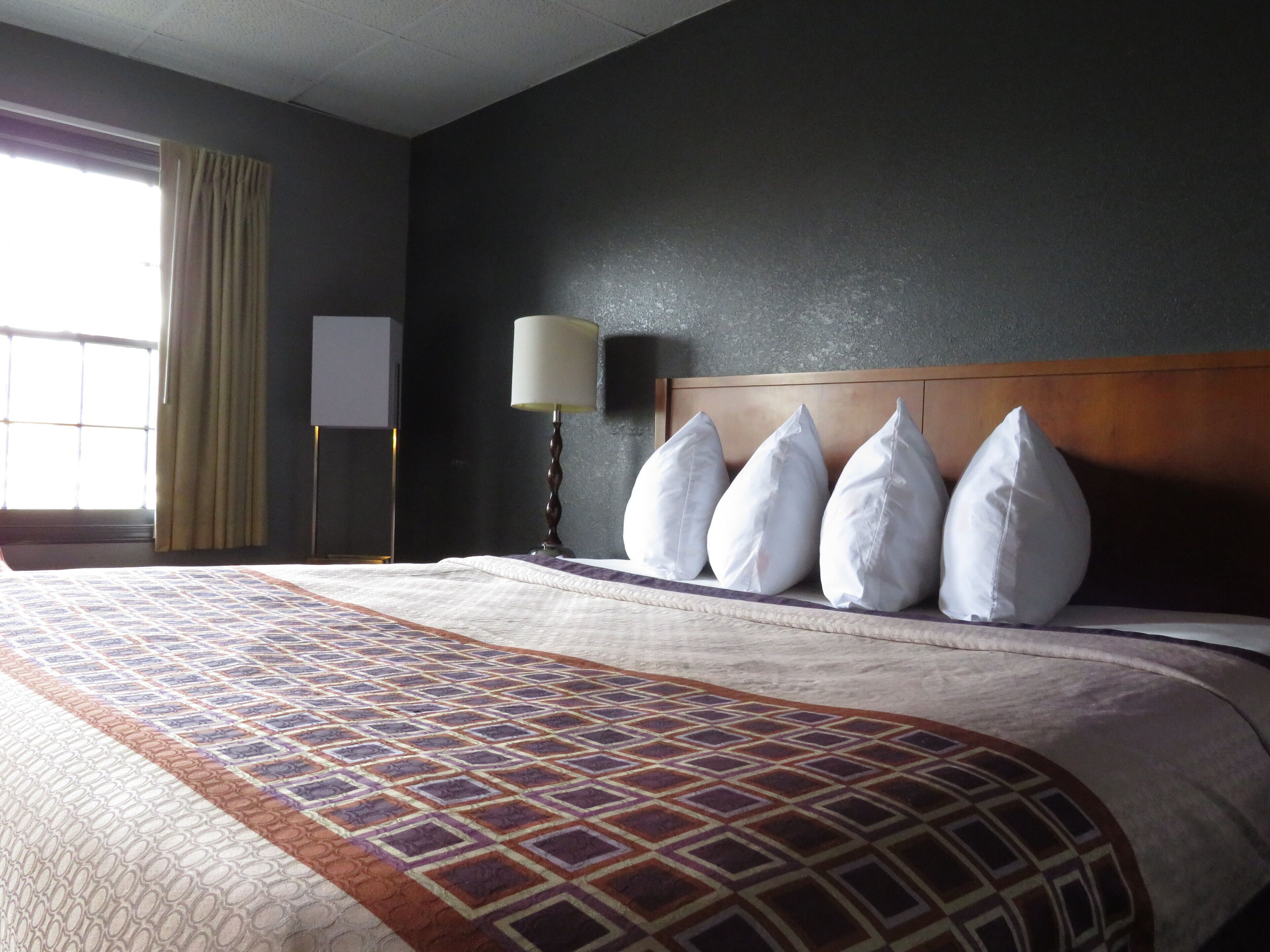 A hotel room with a large bed, a headboard with multiple pillows, a bedside lamp, and a window with curtains.
