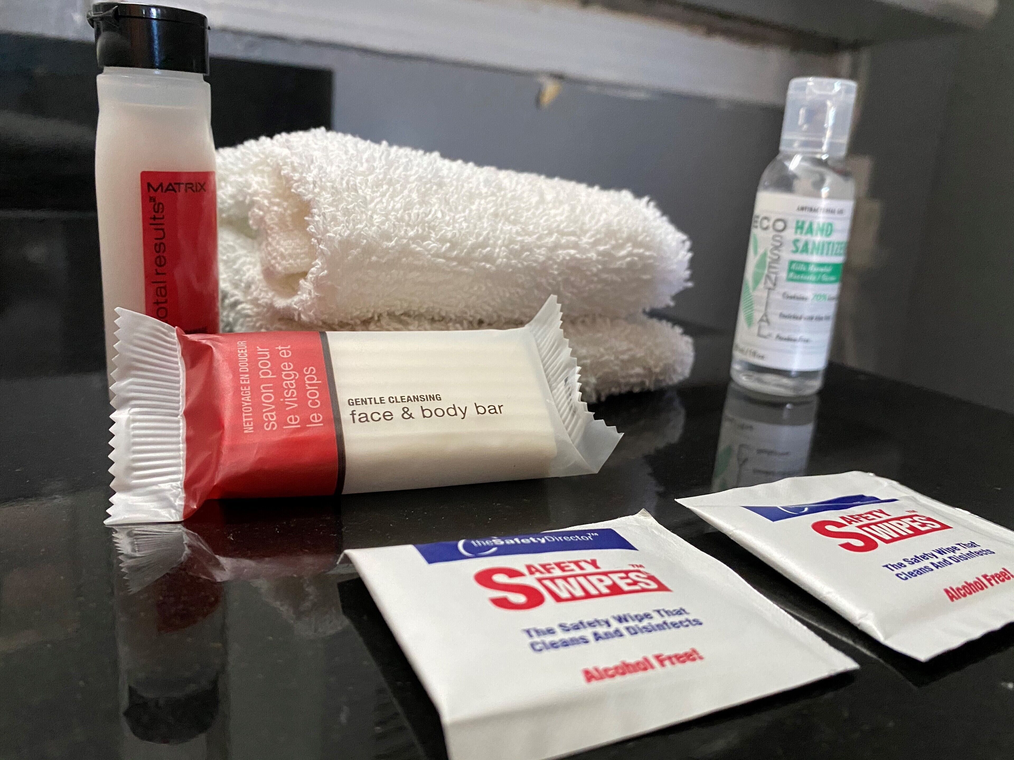 A bathroom counter with a bottle of Matrix Total Refresh, a bar of gentle cleansing face and body bar, a bottle of ECO HAND SANITIZER, and two packets of Safety Swipes.