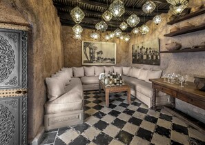 A cozy living room with a checkered floor, a large L-shaped sofa, a wooden coffee table, and a wall-mounted shelf with pottery.