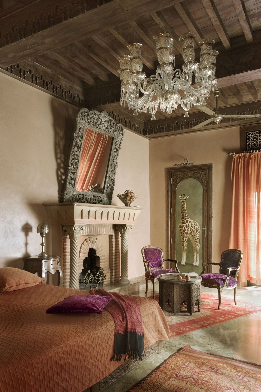 A room with a bed, two chairs, a fireplace, a chandelier, and a giraffe painting.