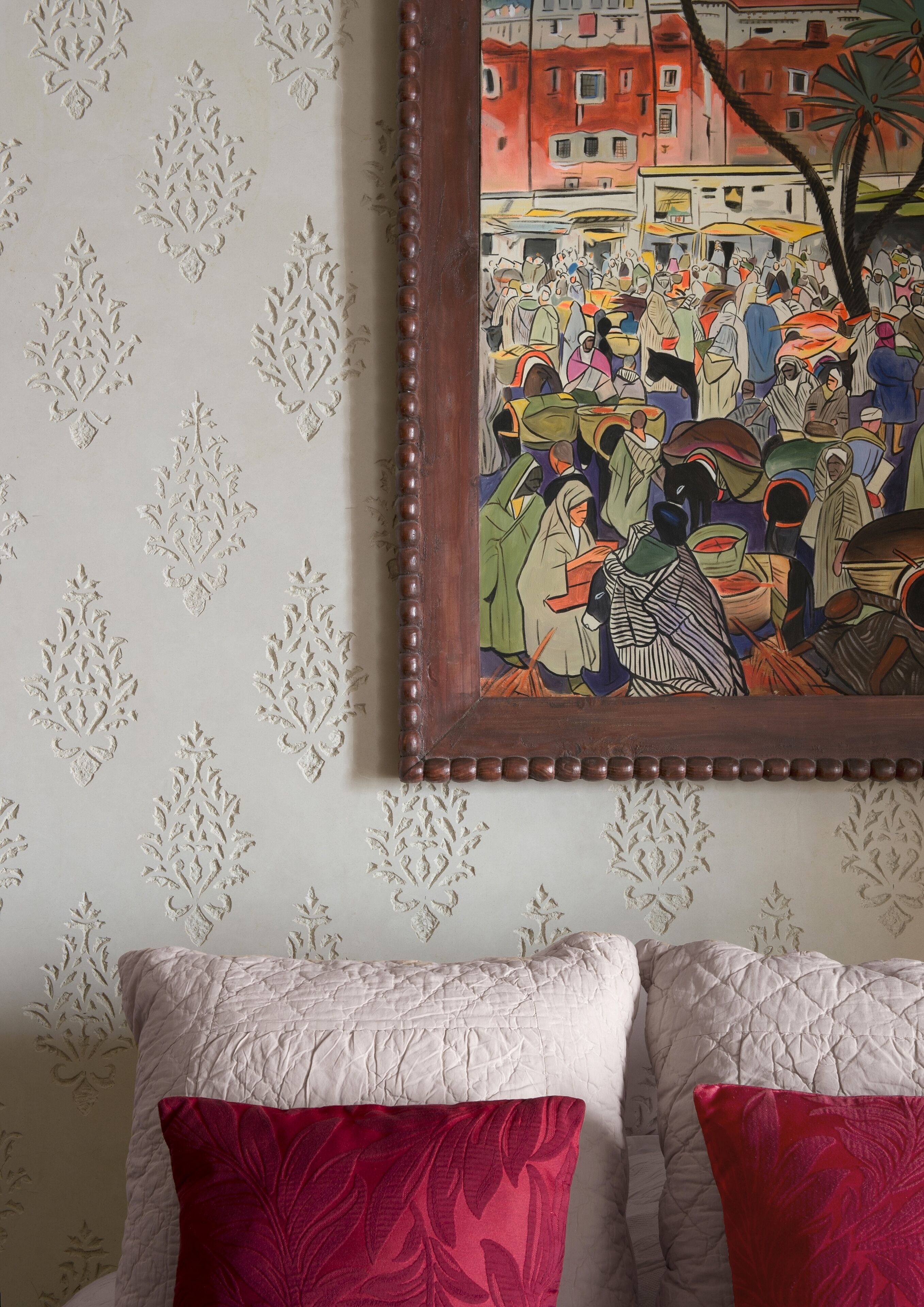 A wall with a framed painting depicting a crowded scene, a patterned wallpaper, and a bed with two pillows.
