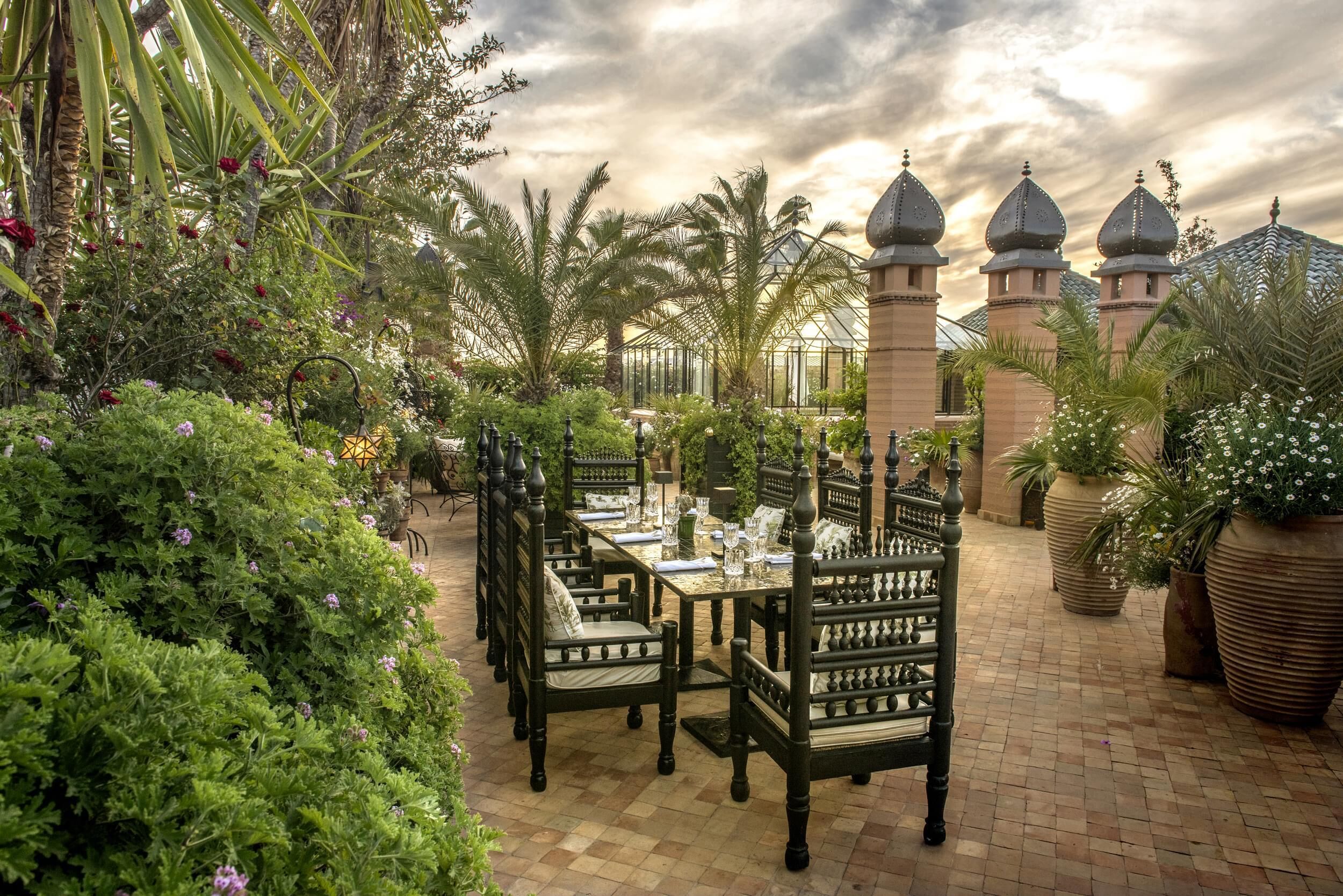 An outdoor dining area with a long table set for two, surrounded by lush greenery and palm trees.