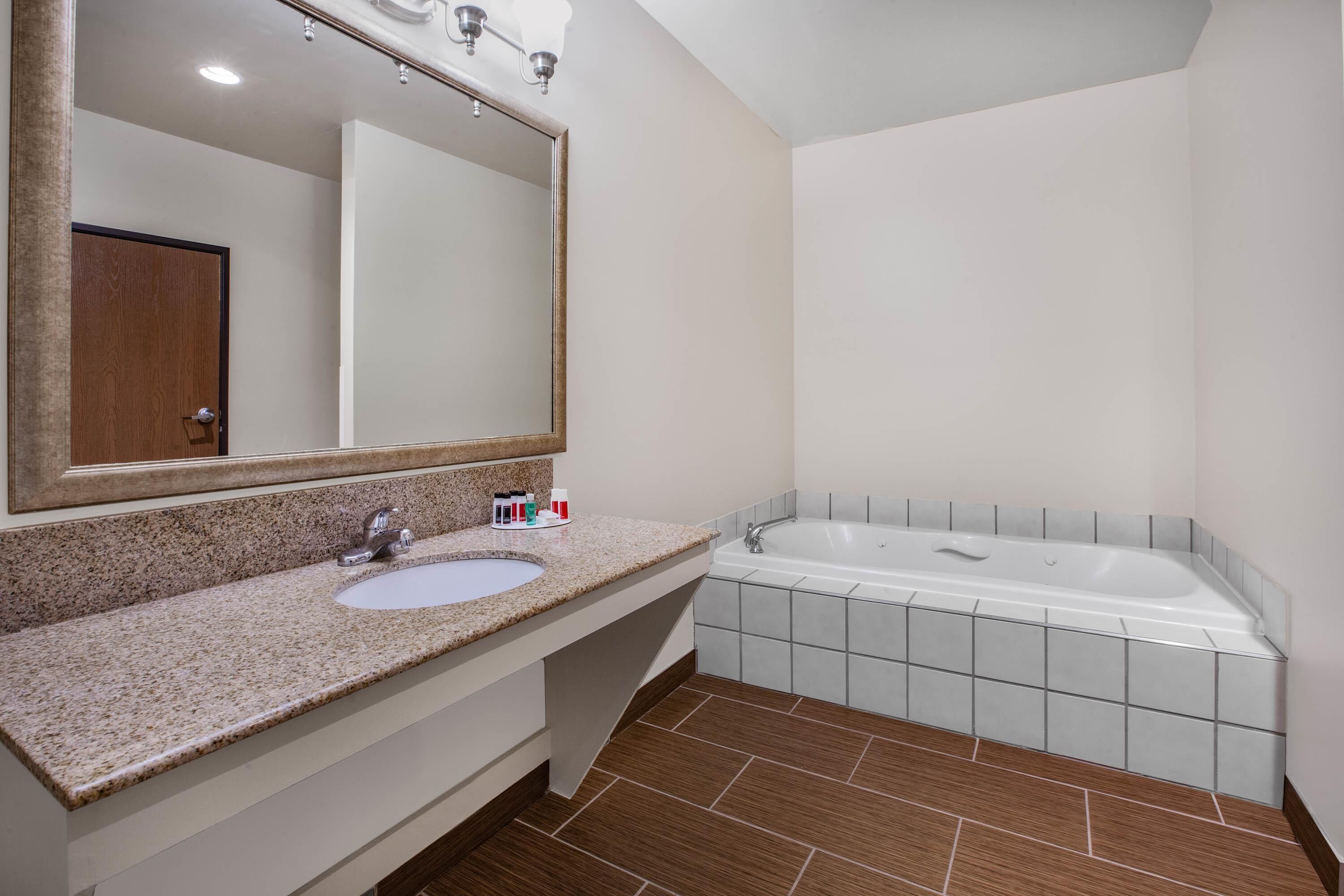 Un baño con lavamanos de encimera de granito, un espejo grande, una bañera blanca y suelo de baldosas marrones.
