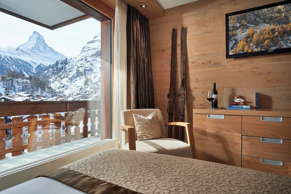 A bedroom with a large window, wooden furniture, a ski rack, and a painting of a mountain landscape.