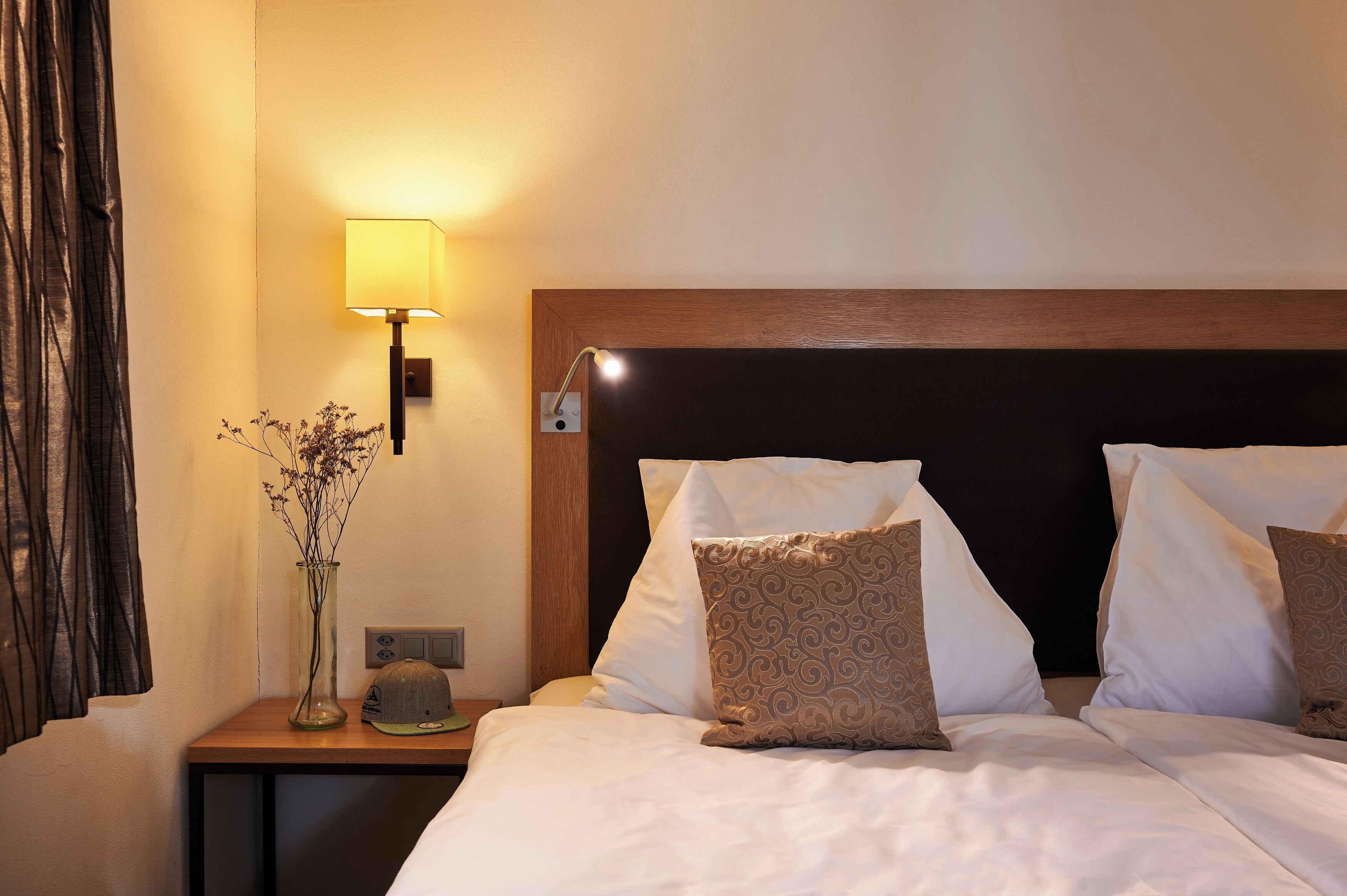 A hotel room with a bed, pillows, a nightstand, a lamp, and a vase with dried flowers.