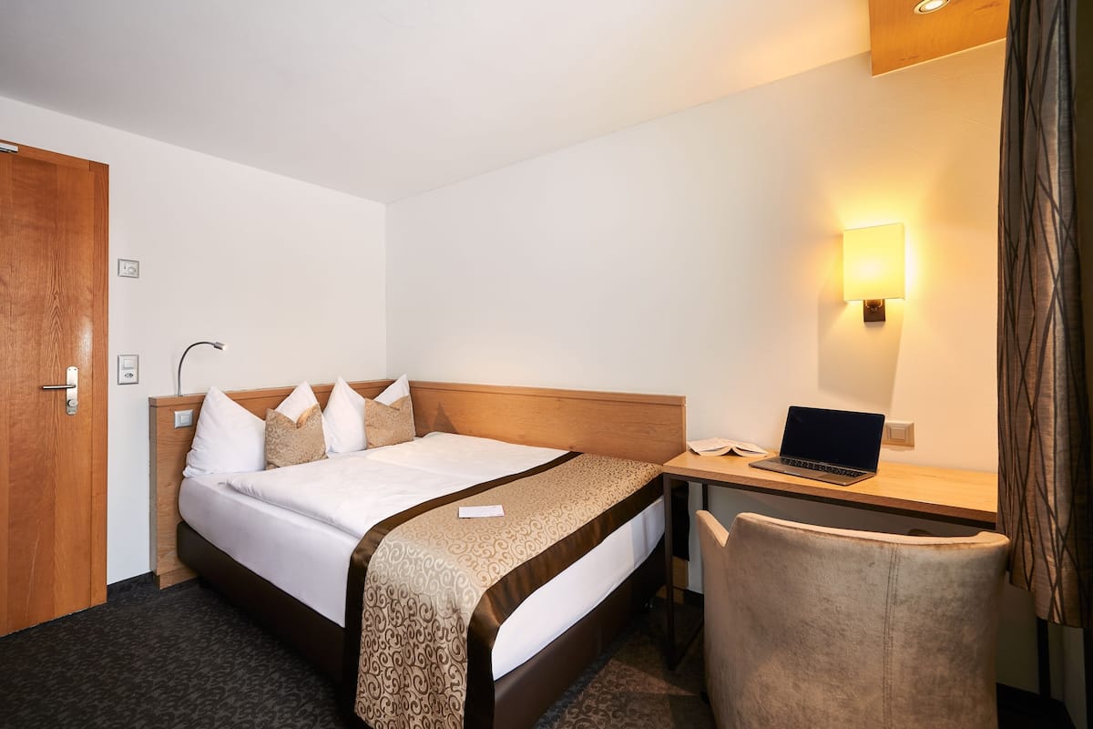 A hotel room with a bed, a desk with a laptop, a chair, a door, and a wall-mounted light.