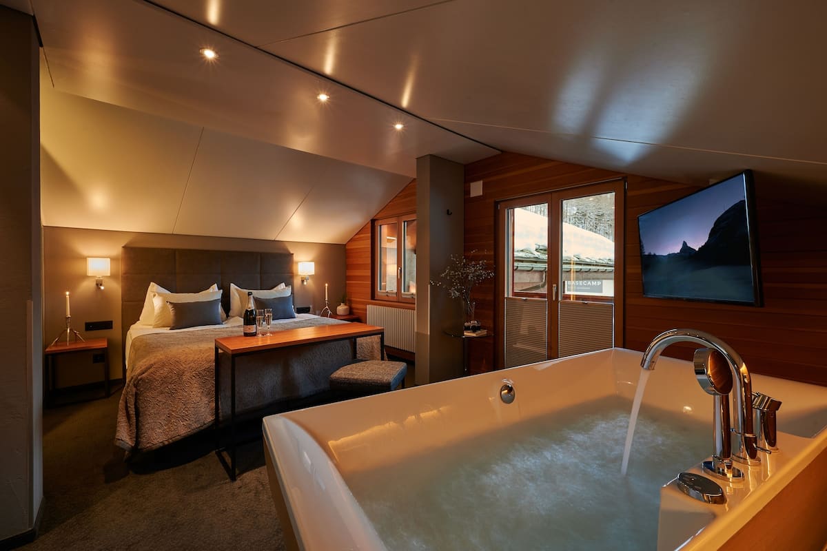 A modern bedroom with a large bathtub, a bed with pillows, a bedside table with a bottle, and a TV on the wall.