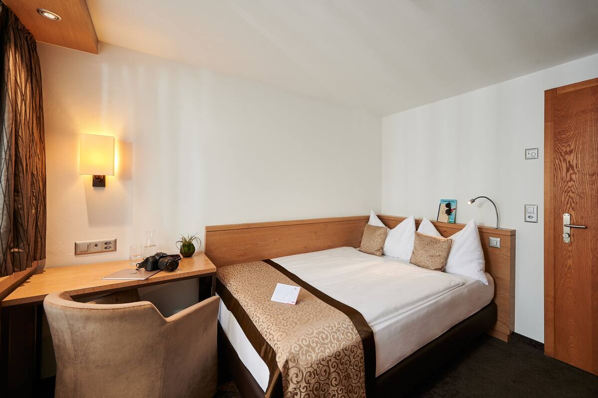 A hotel room with a bed, a desk, a chair, a nightstand, a lamp, and a wall-mounted light.