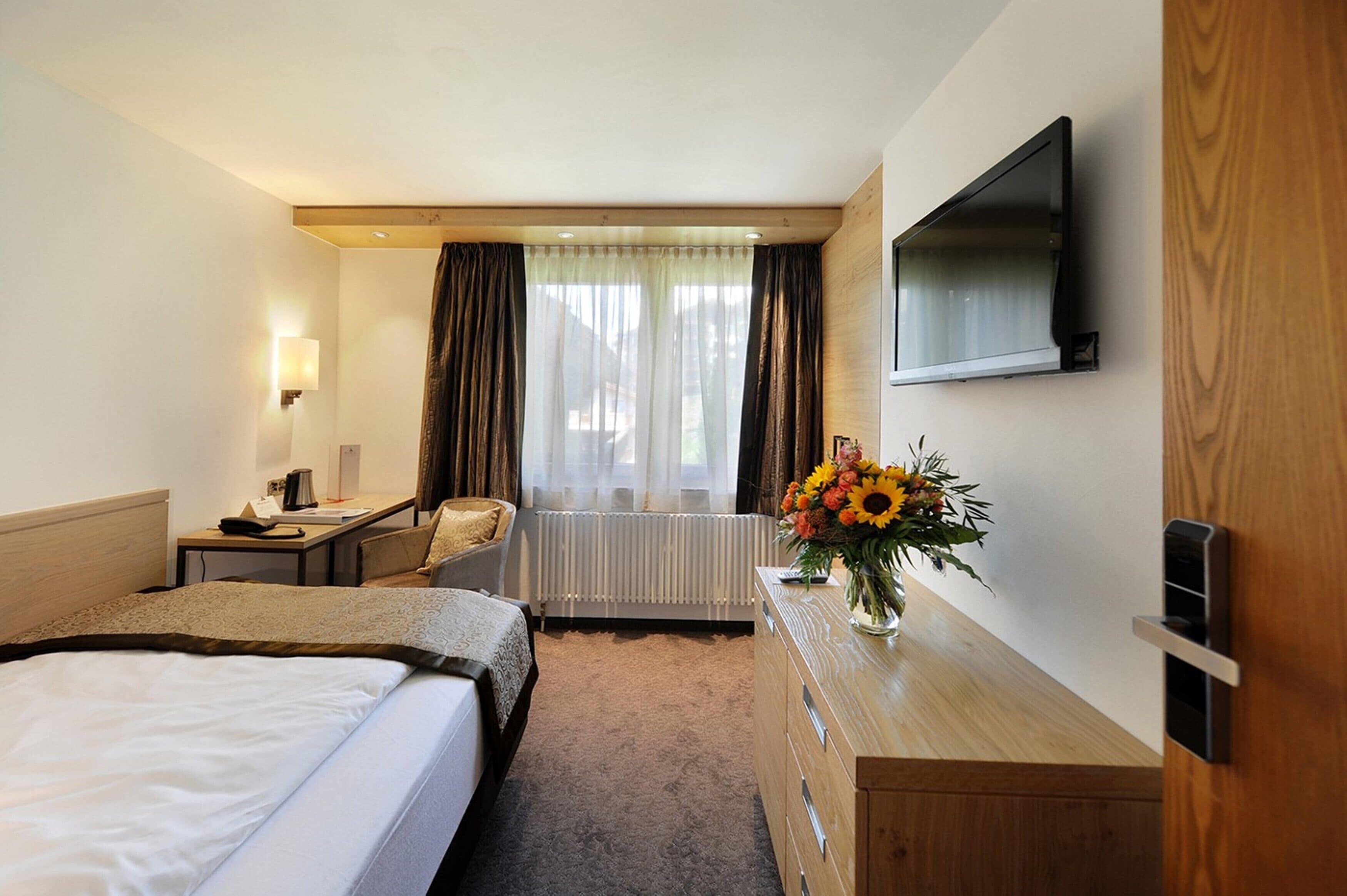 A hotel room with a bed, a desk, a chair, a dresser, a TV mounted on the wall, and a window with curtains.