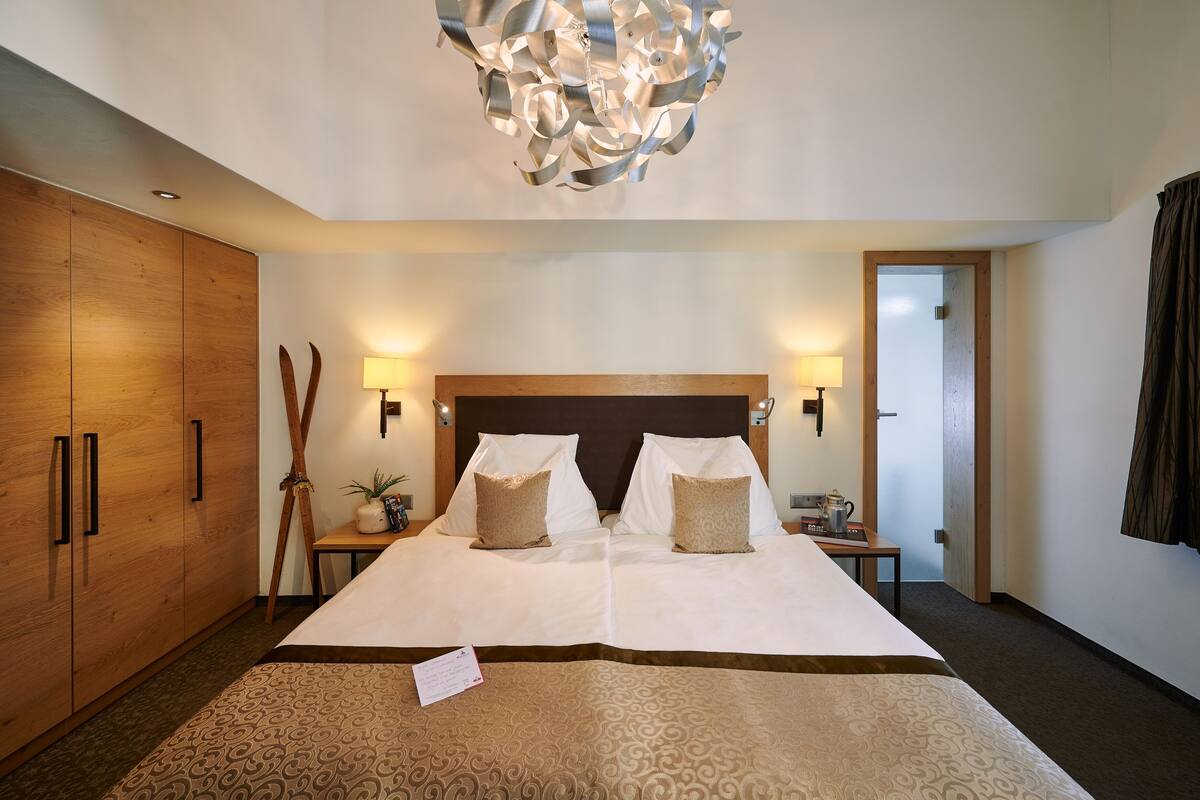 A hotel room with a large bed, wooden wardrobe, two bedside tables with lamps, and a modern chandelier.
