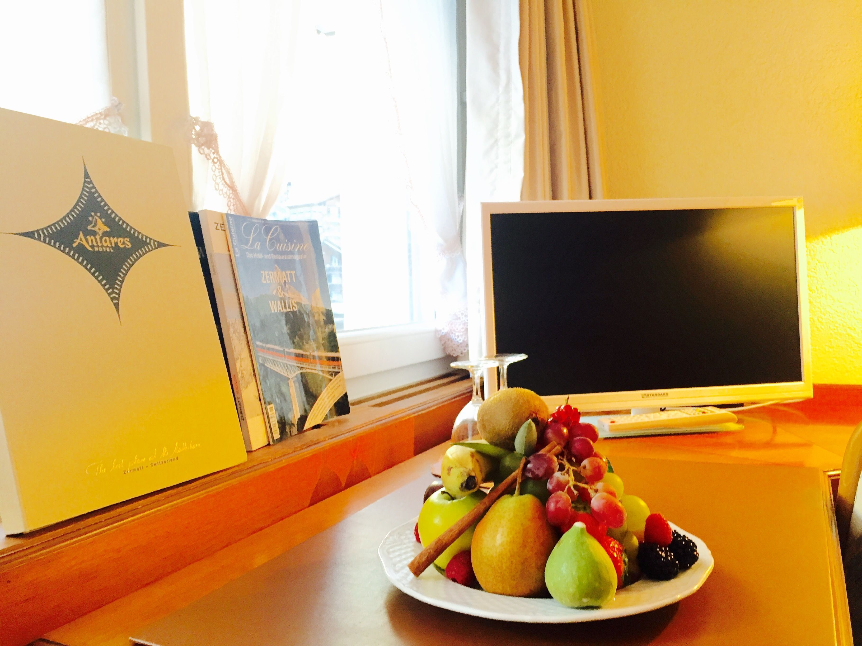 Ein Hotelzimmer mit einem Holztisch, Obstteller, einem Buch und einem Fernseher auf einem Schrank.
