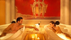 A couple in a hot tub with a painting of nude figures on the wall behind them.