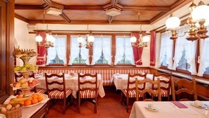 A dining area with wooden tables and chairs, a buffet with fruits and pastries, and a view of a building through large windows. - Hotel Astoria (Zermatt)