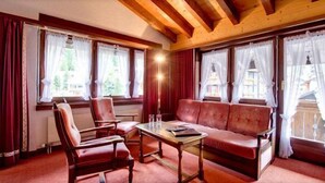A room with a wooden ceiling, a red sofa, two armchairs, a table, and large windows with curtains. - Hotel Astoria (Zermatt)