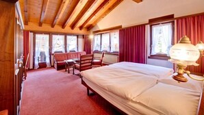 A hotel room with a wooden ceiling, a large bed, a desk, a chair, and a window with curtains. - Hotel Astoria (Zermatt)