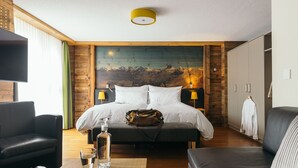Habitació d'hotel amb un llit gran, un panell de fusta al capçal, un quadre, una televisió, un sofà de cuir, una taula amb un got i un llibre, i un armari.