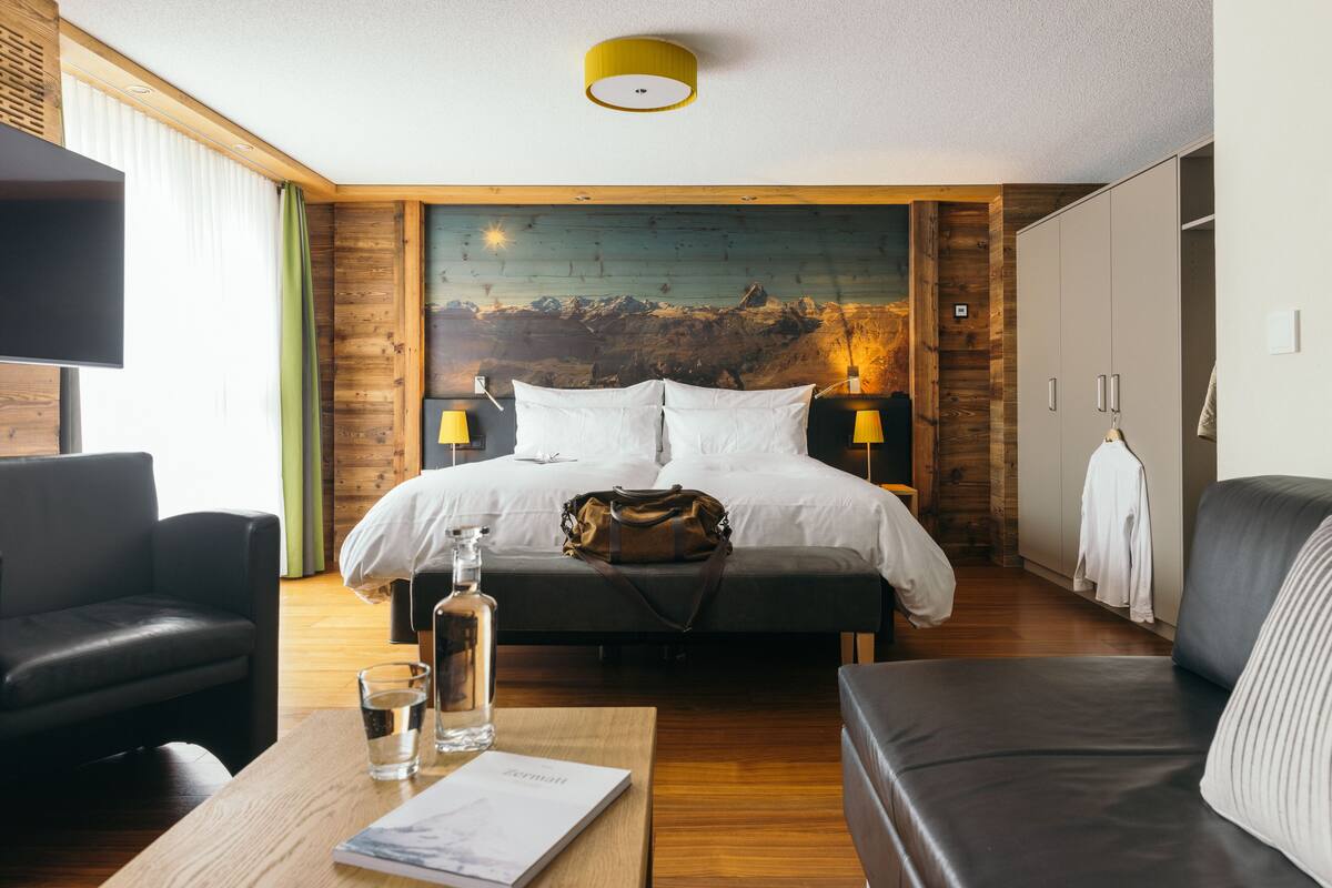 A hotel room with a large bed, a wooden headboard, a painting, a TV, a leather sofa, a table with a glass and a book, and a wardrobe.