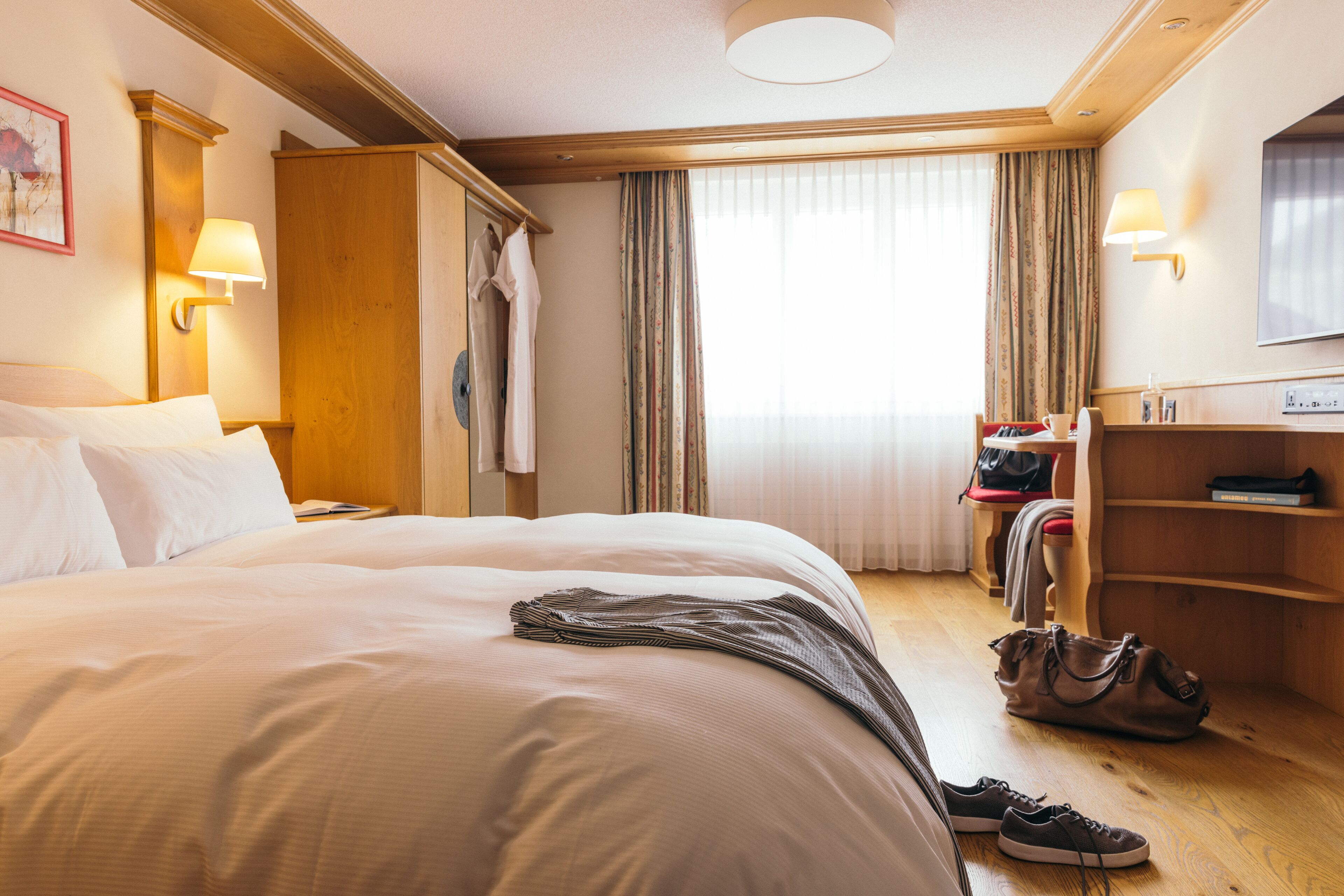 A hotel room with a large bed, a wooden wardrobe, a desk with a chair, a television, and a window with curtains.