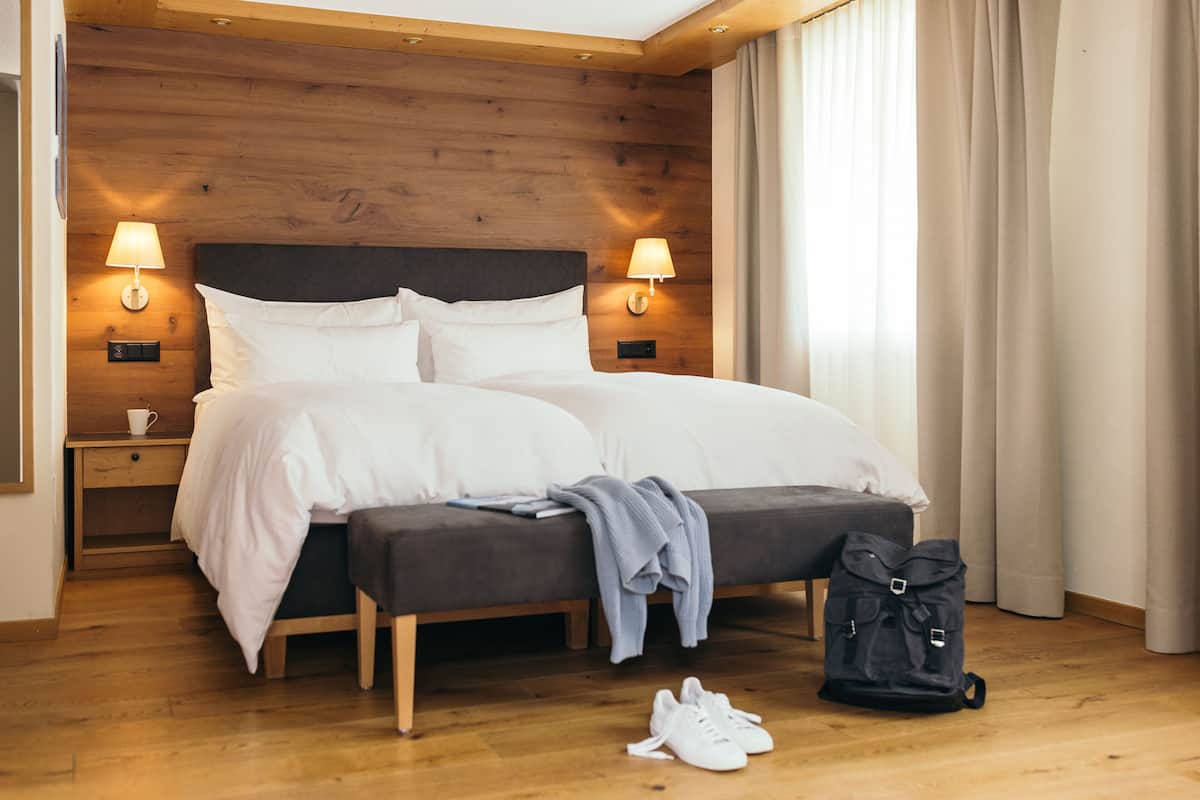 A hotel room with a wooden headboard, two bedside lamps, a bed with white bedding, a bench, a backpack, and a pair of sneakers on the floor.