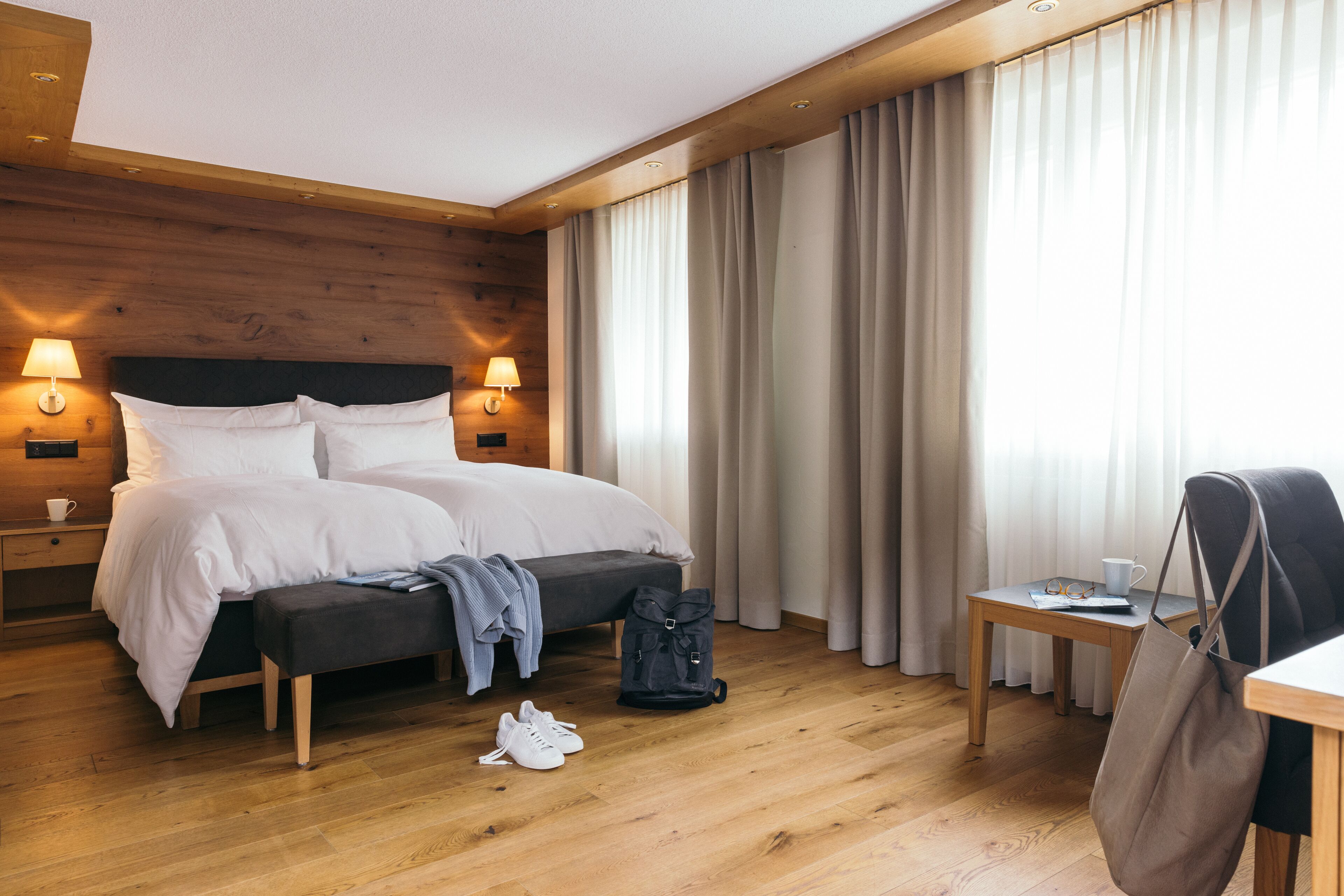 A hotel room with a wooden headboard, two bedside lamps, a bed with white linens, a small table with a cup and glasses, a bench, and a backpack on the floor.