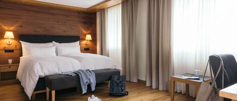 A hotel room with a wooden headboard, two bedside lamps, a bed with white linens, a small table with a cup and glasses, a bench, and a backpack on the floor.