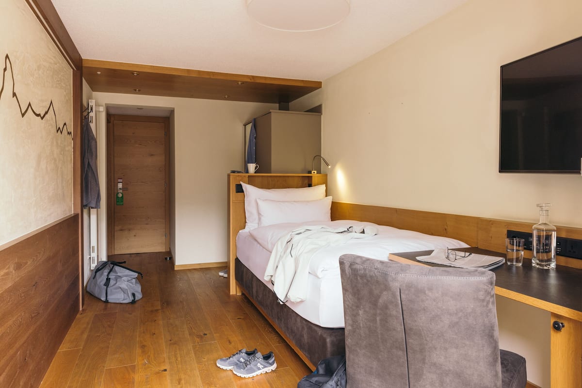 A hotel room with a wooden floor, a bed with white linens, a desk with a mirror, a chair, and a TV mounted on the wall.