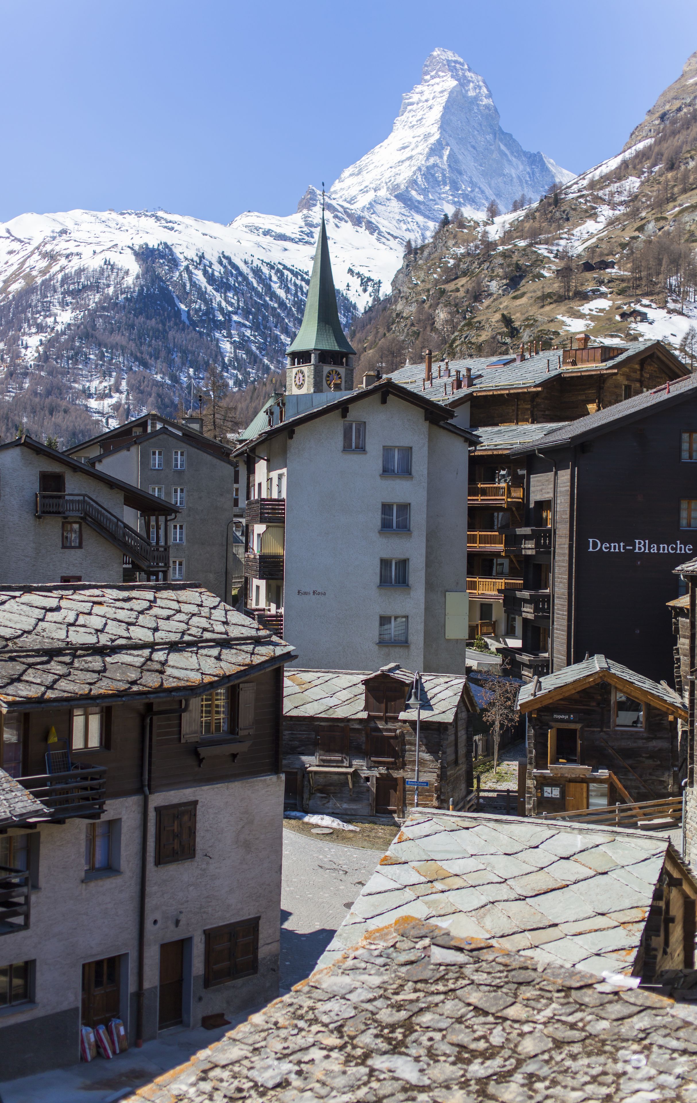 A traditional European village with stone buildings, a church tower, and a snow-covered mountain in the background.
