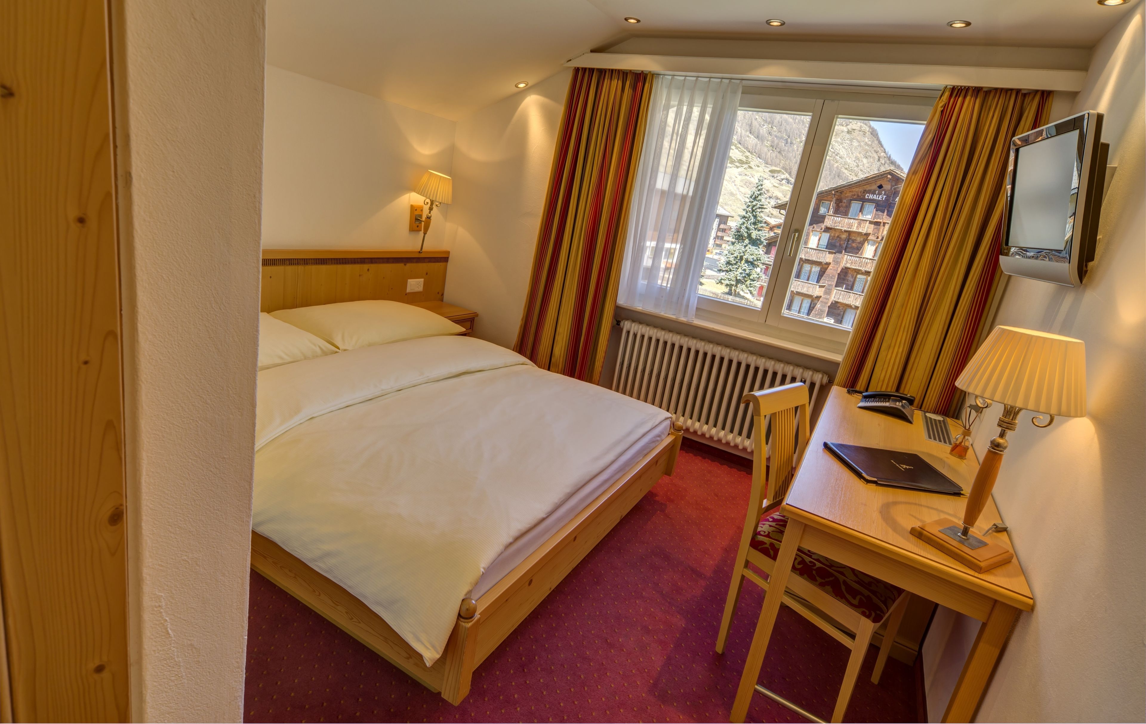 A hotel room with a bed, a desk, a chair, a lamp, a TV, and a window with curtains.