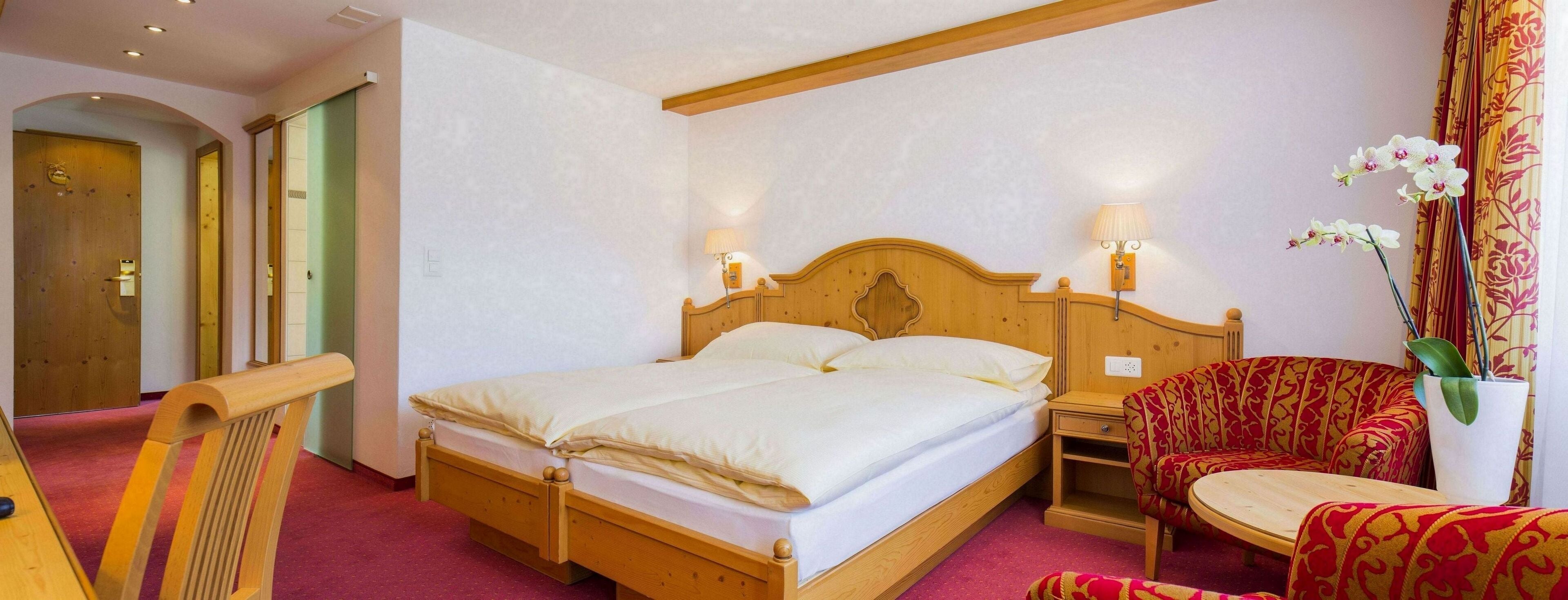 A hotel room with a wooden bed, bedside lamps, a red patterned armchair, and a small table.