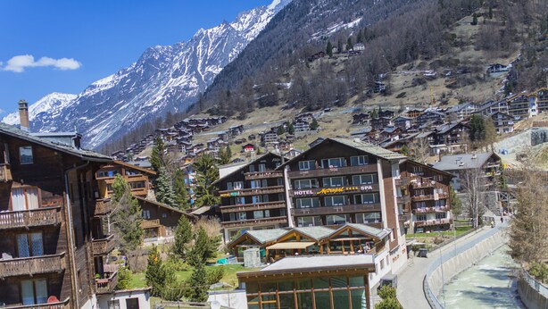 A mountain resort with wooden buildings, a river, and snow-capped mountains in the background. - Hotel Excelsior (Zermatt)