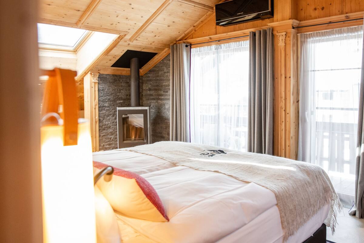 A bedroom with a bed, a stone wall, a fireplace, and large windows with curtains.