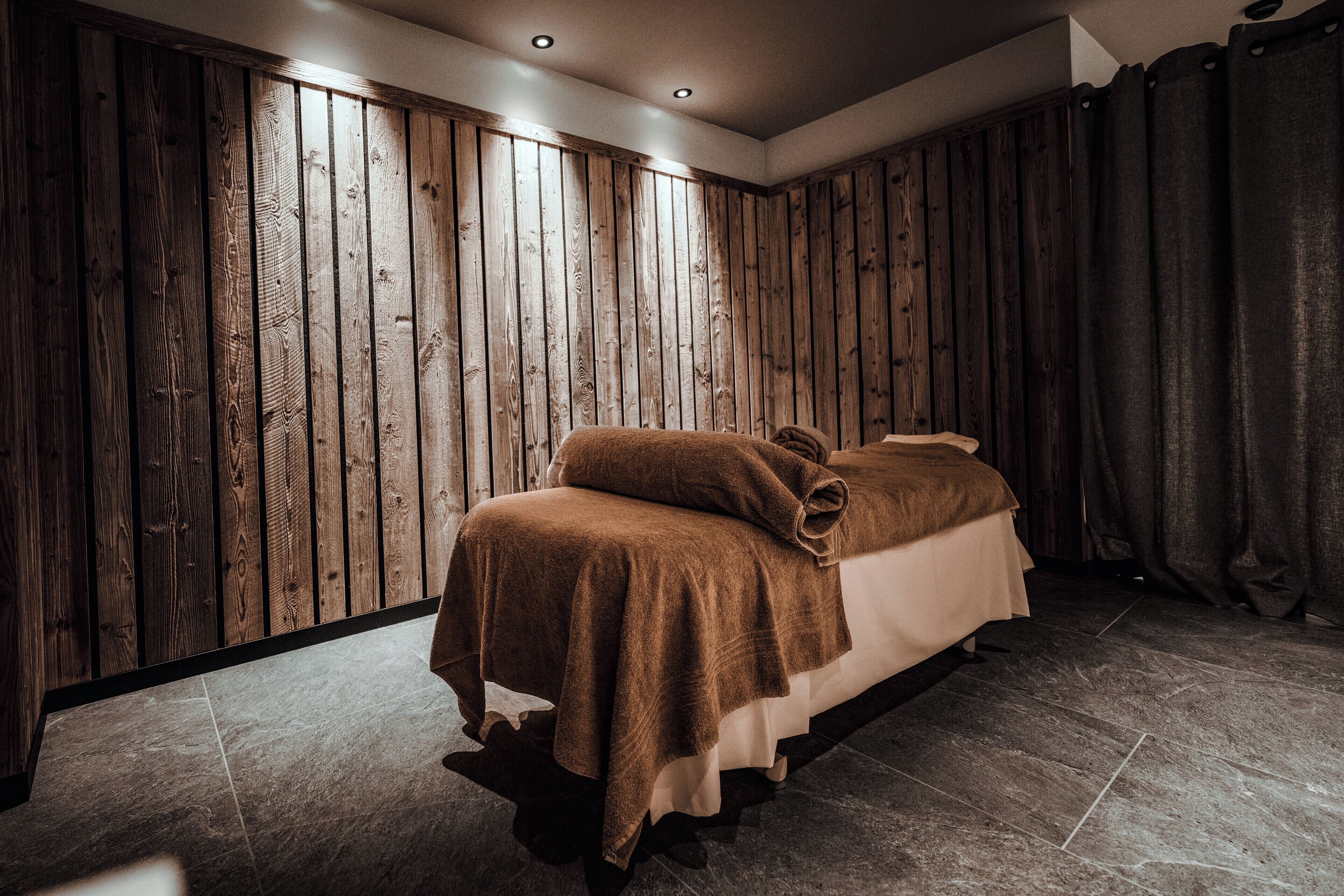 A room with a wooden panel wall, a bed with a brown blanket, and a tiled floor.