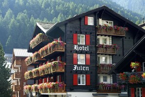 Un hotel a più piani con balconi decorati da fioriere e uno sfondo montuoso, con un cartello che riporta 'Hotel Julen'.