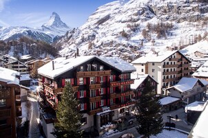 Un hotel de estilo arquitectónico alpino tradicional, con varios pisos y balcones, con montañas nevadas y cielo despejado de fondo.