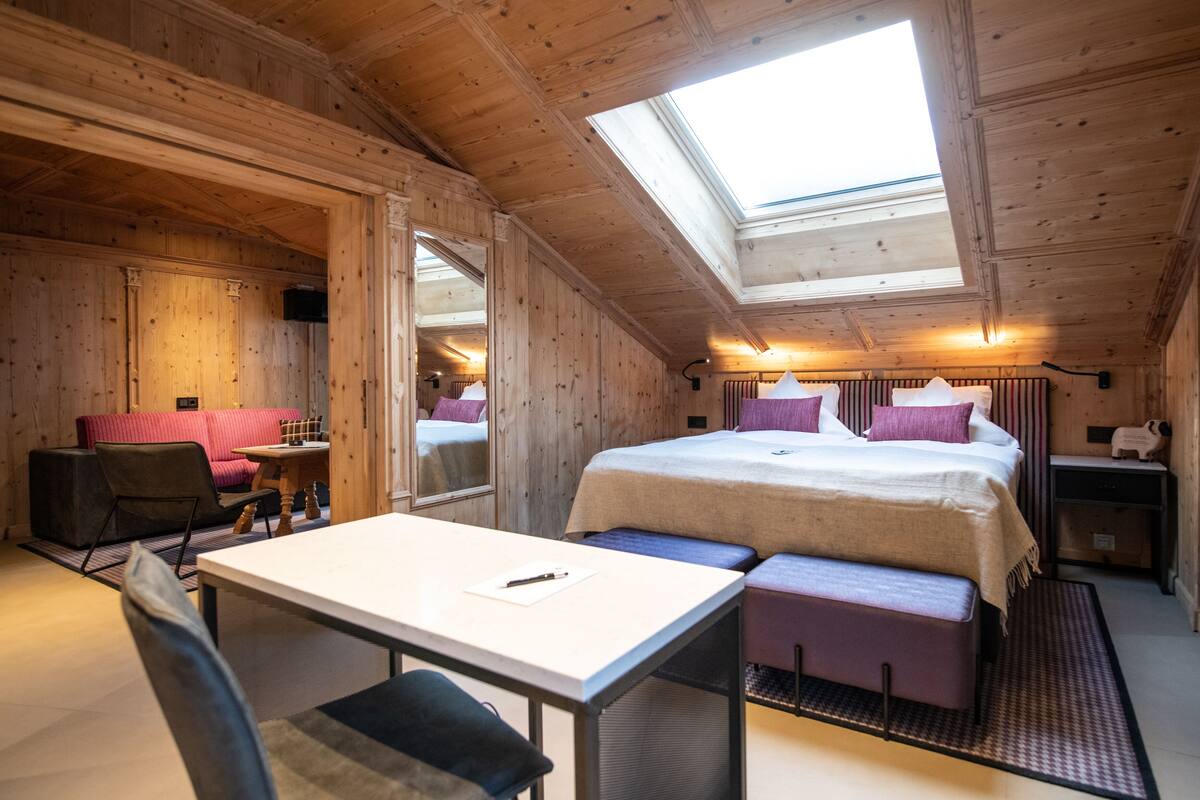 A cozy room with a bed, a desk, and a chair. There is a skylight above the bed, and the room has wooden walls and ceiling.