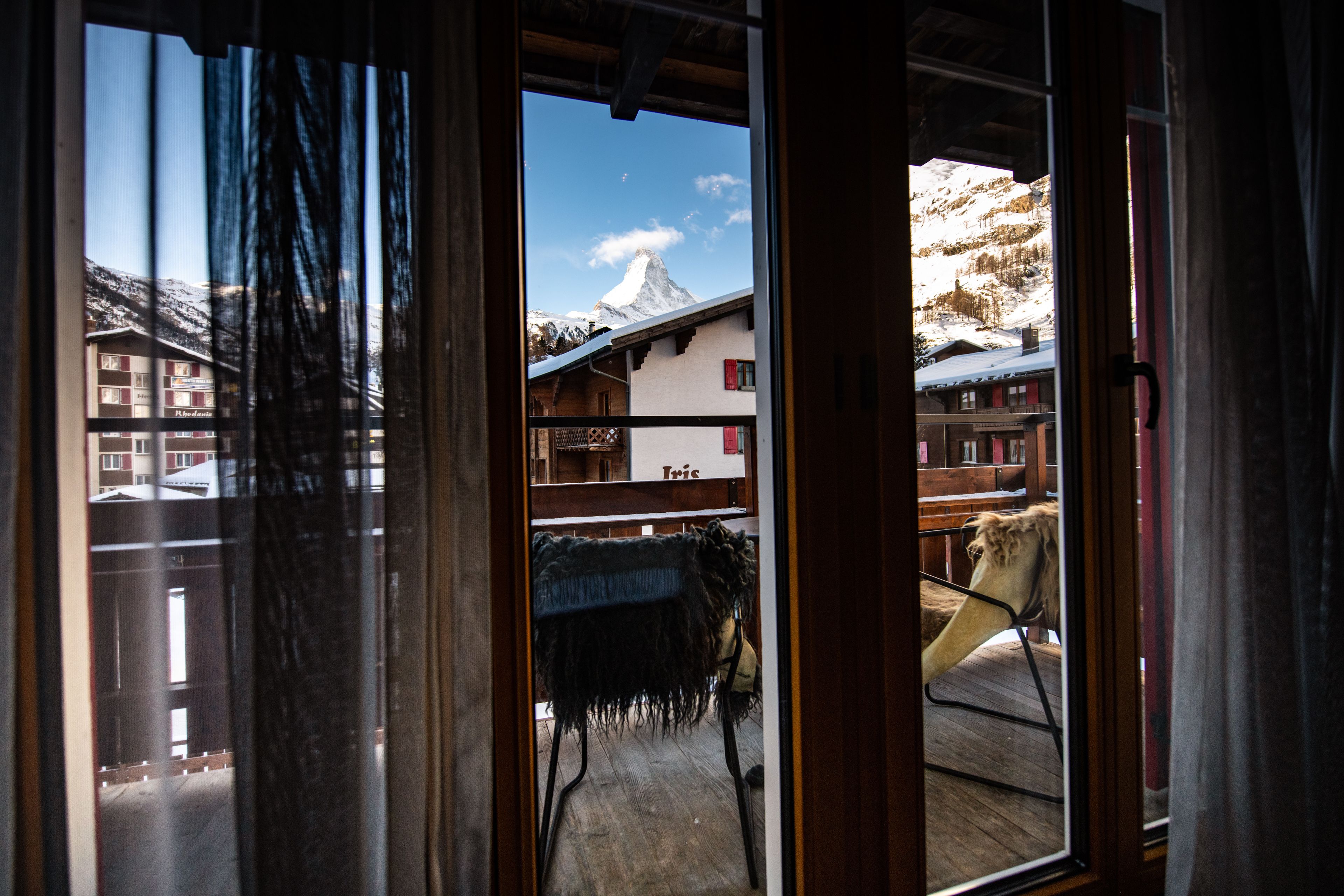 A view through a window of a building with a wooden deck, a chair, and a mountain in the background.
