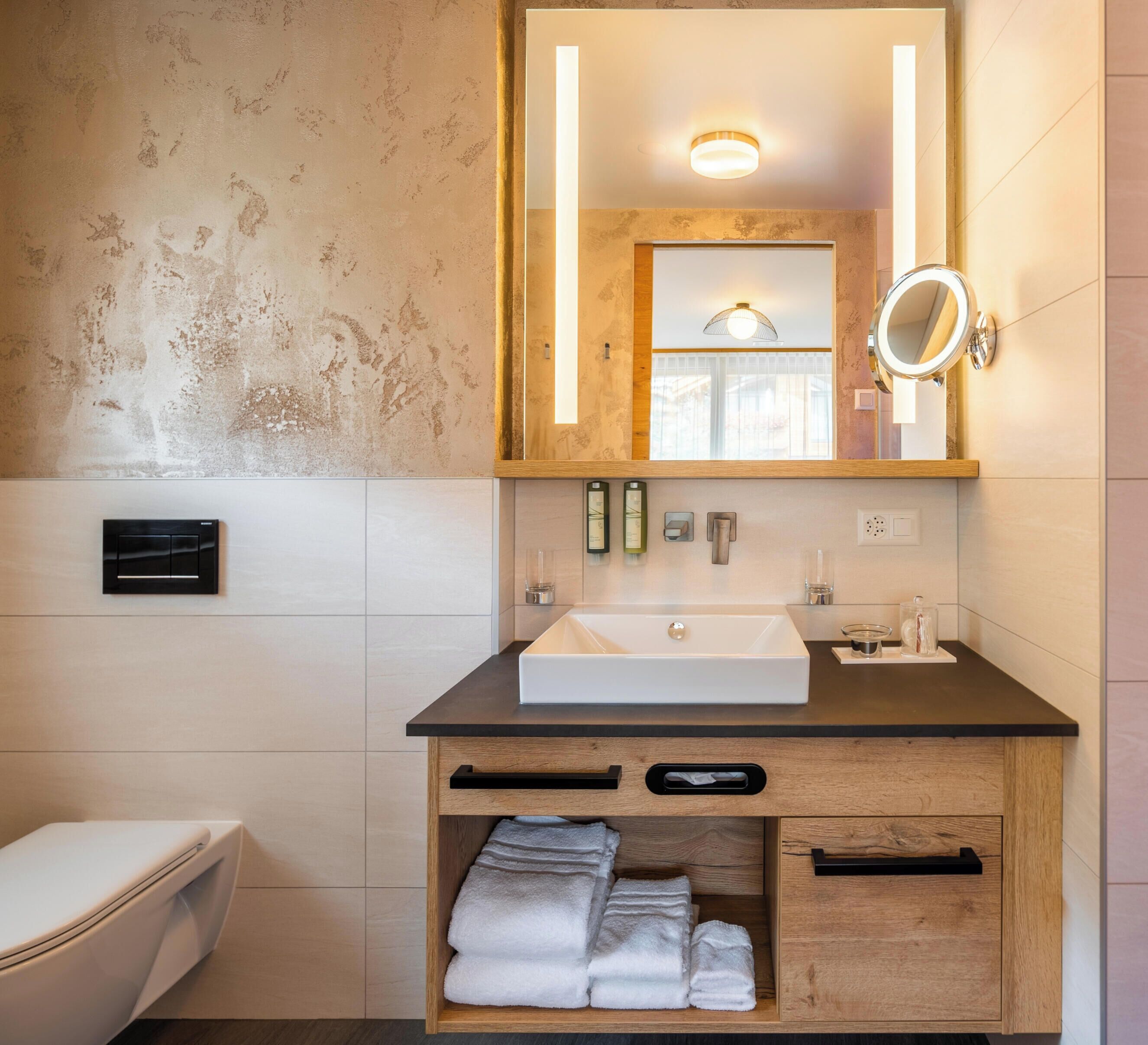 Ein Badezimmer mit weißem Waschbecken, Holzkommode, einem Spiegel mit Beleuchtung und einem Handtuchhalter.