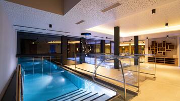 A modern indoor swimming pool with clear glass walls, a metal staircase, and contemporary lighting fixtures.