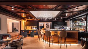 A modern bar area with a curved counter, bar stools, and a variety of bottles. There are seating areas with chairs and tables, and a ceiling with a unique light fixture. - Resort La Ginabelle (Zermatt)